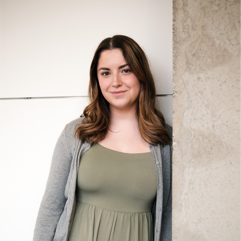 Portrait d’Andréanne Lacasse portant une robe verte et un cardigan gris devant un mur blanc.