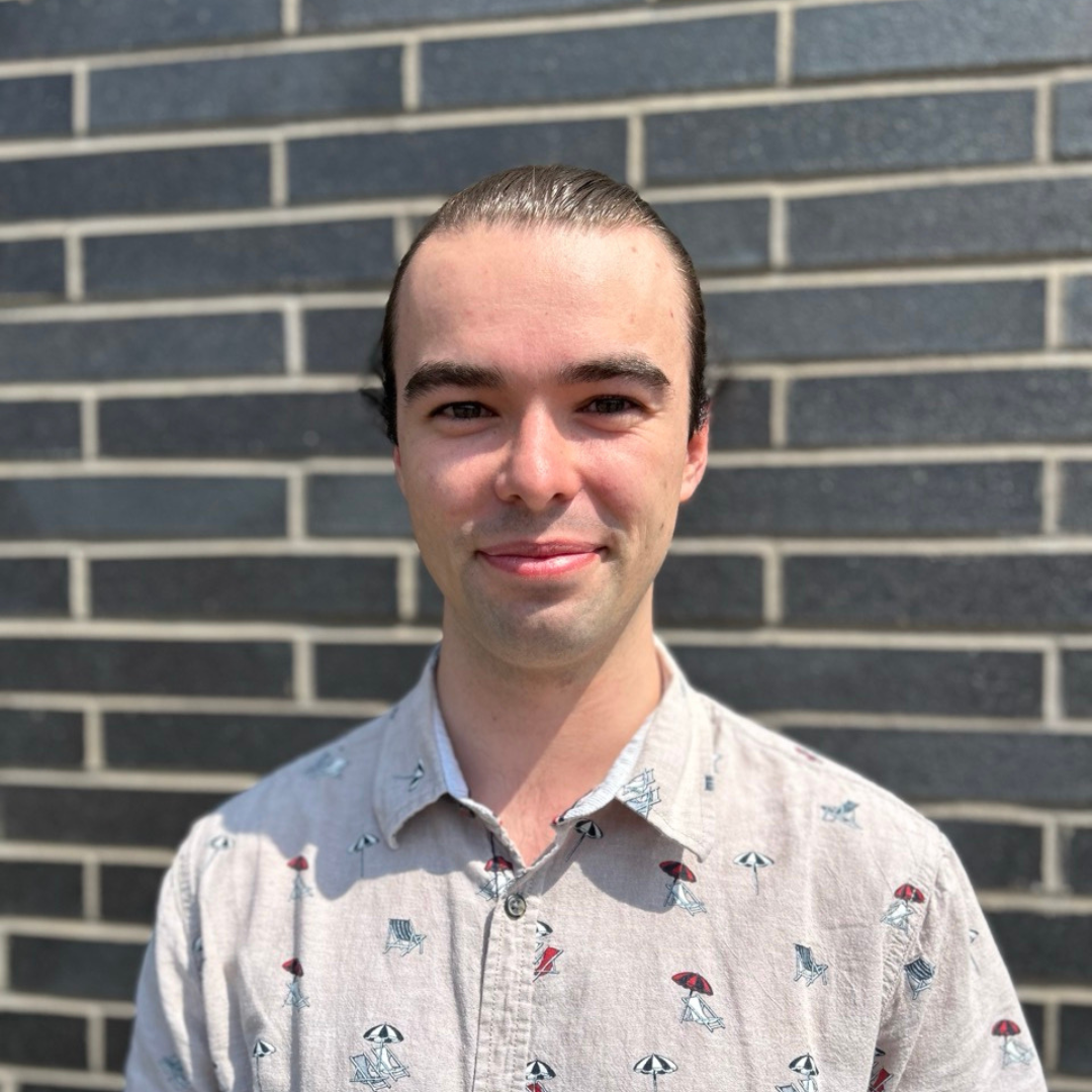 Portrait de Gabriel Olivier-Fortier qui porte une chemise grise avec des parasols devant un mur de briques.